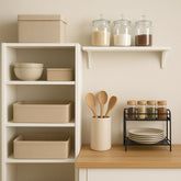 A small, neatly organized kitchen corner featuring open white shelving with beige storage bins, baskets, and bowls. A wooden countertop holds a utensil holder and black metal racks storing
